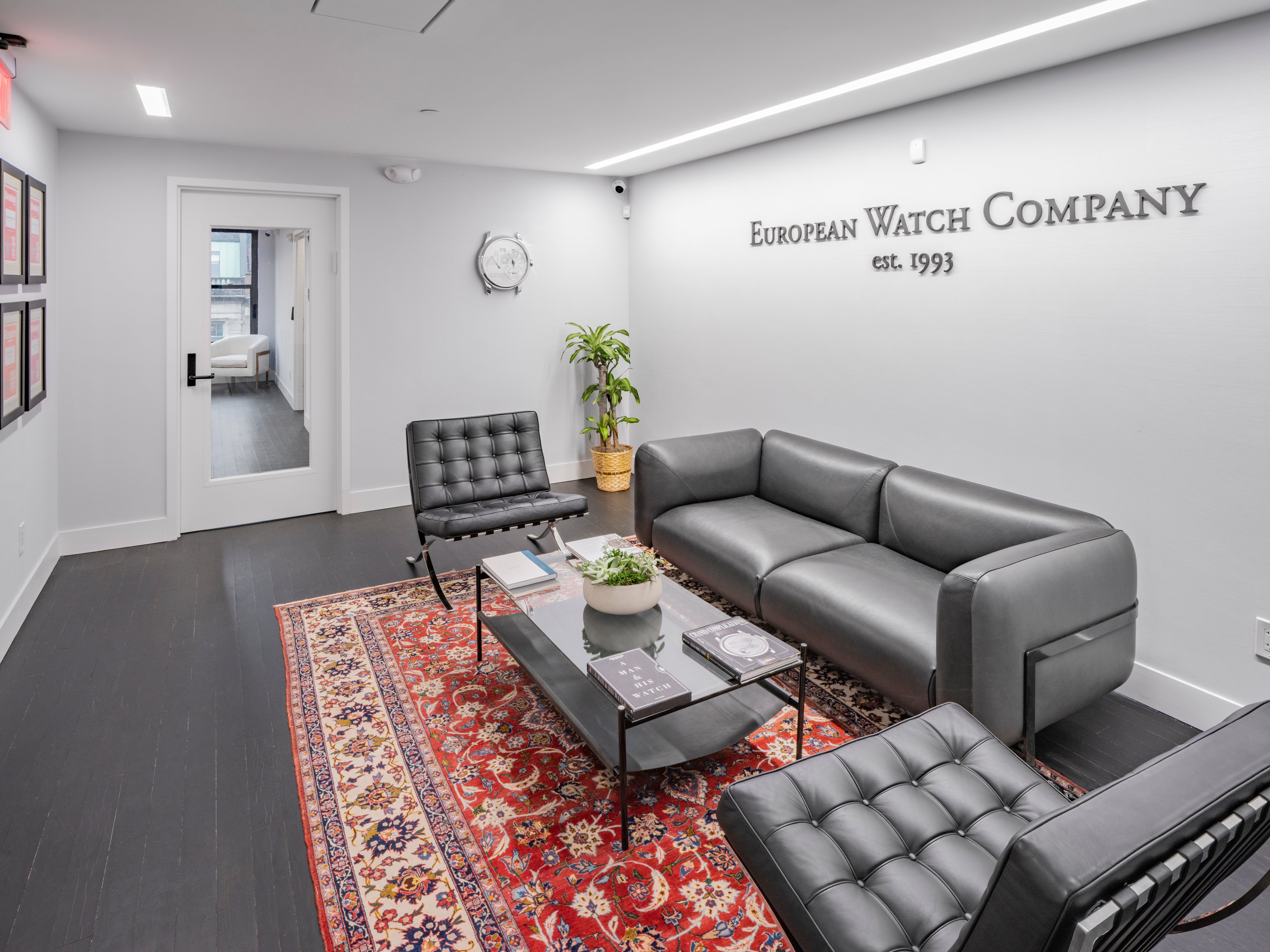 Interior of European Watch Company, lounge area, 3 love seats, oriental area rug with glass coffee table with  plants, books, with "European Watch Company est. 1993" on the wall behind center love seat, plants in the corner, angle shows clock, wall decor, and glass door as entrance